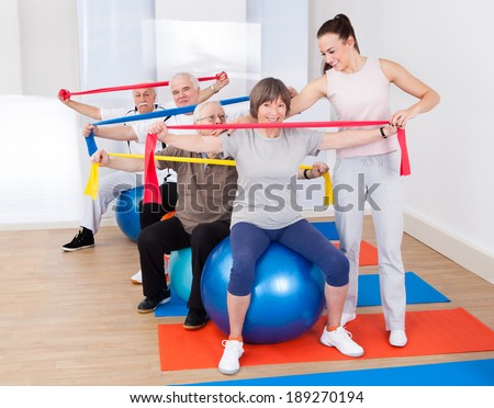Young female trainer assisting senior people with resistance bands sitting on fitness balls at gym