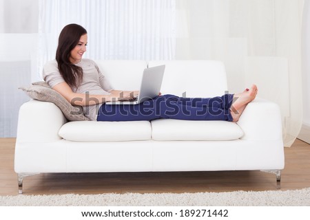 Smiling young woman using laptop while relaxing on sofa at home