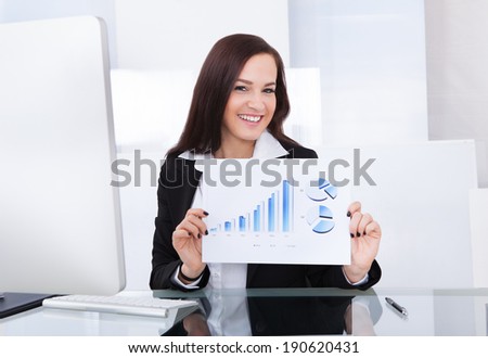 Portrait of happy confident businesswoman showing progress chart at desk in office