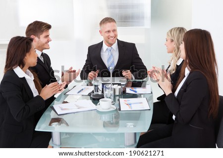 Happy businesspeople clapping for colleague after presentation at desk in office