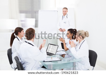 Team of young doctors clapping for colleague after presentation at desk in clinic