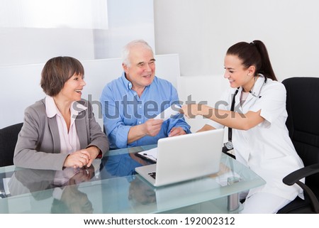 Smiling female doctor giving prescription paper to senior couple at desk in clinic