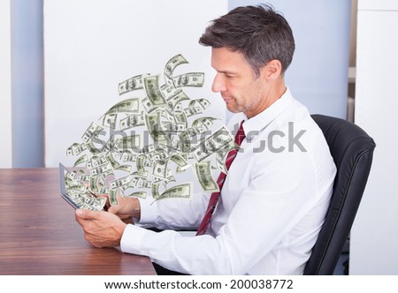Side view of mature businessman looking at money coming out of digital tablet in office