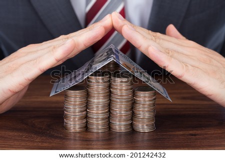 Midsection of businessman sheltering coins and banknote in house shape on table