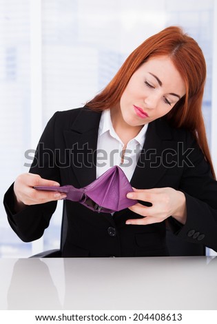 Upset young businesswoman looking at empty wallet while sitting at office desk