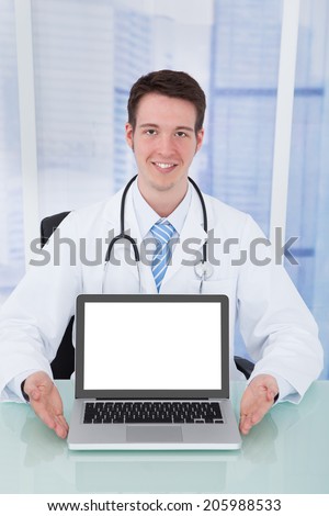 Portrait of confident young male doctor presenting laptop with blank screen at desk in hospital