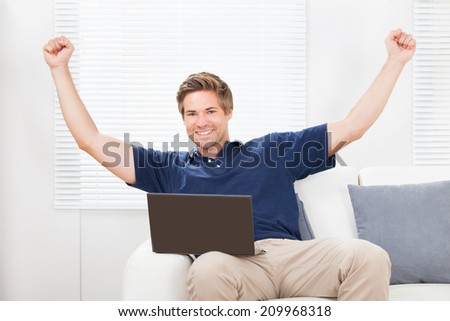 Portrait of excited man with laptop raising hands on sofa at home