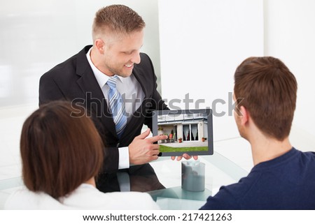 Smiling advisor showing house picture to couple on tablet at office desk