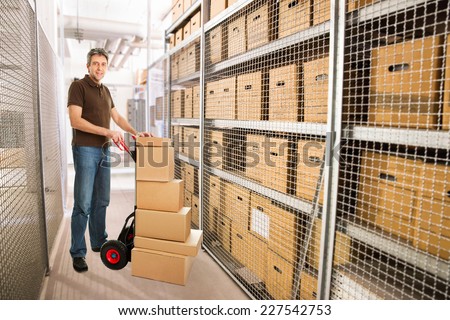 Portrait of delivery man with stacked cardboard boxes on hand truck at warehouse