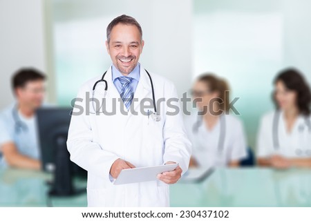 Portrait of happy mature doctor holding digital tablet against team in meeting room