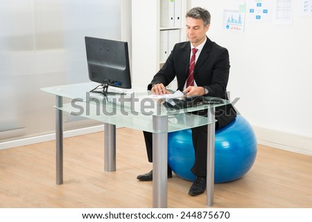 Businessman Writing On Paper Sitting On Pilates Ball In Office