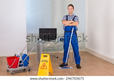 Portrait Of Happy Confident Male Janitor With Cleaning Equipments And Wet Floor Sign In Office