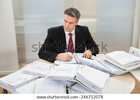 Mature Businessman Working At Office With Heap Of Folders Around Him