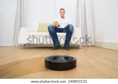 Happy Man Operating Robotic Vacuum Cleaner With Remote Control At Home