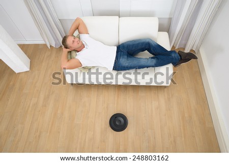 Man Relaxing On Couch With Robotic Vacuum Cleaner On Hardwood Floor
