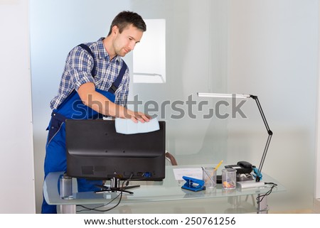 Portrait Of Happy Male Janitor Cleaning Computer On Office Desk