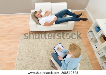 Patient Lying On Couch In Front Of Psychiatrist Writing On Clipboard