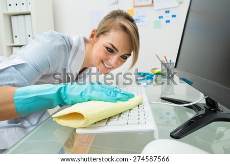 Portrait Of Young Happy Maid Cleaning Keyboard At Desk In Office