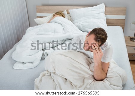 Depressed Man Sitting On Bed While Woman Sleeping In Bedroom