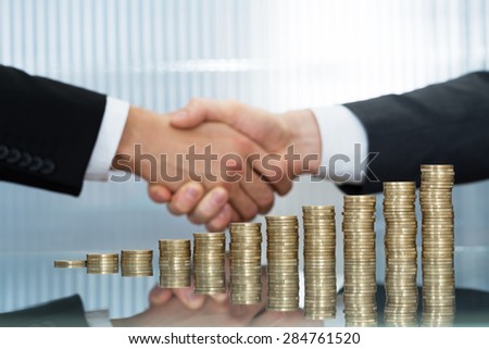Close-up Of Businesspeople Shaking Hand In Front Of Stacked Coins
