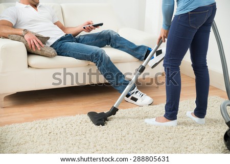 Close-up Of A Woman Cleaning Carpet In Front Of Man Sitting On Couch