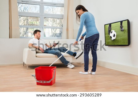 Woman Cleaning Floor In Front Of Man Watching Football Match On Television