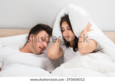 Irritated Woman Covering Her Ears With Pillow While Man Snoring On Bed At Home