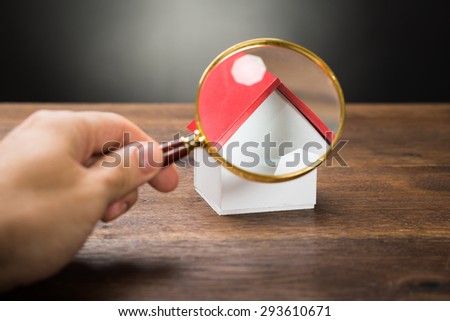 Close-up Of Person Hand Holding Magnifying Glass Over A Miniature House On The Table