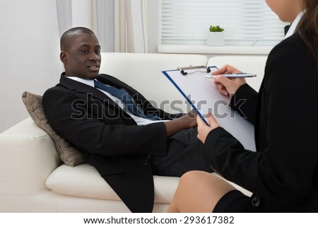 African Patient Sitting On Couch And Female Psychiatrist Writing On Clipboard