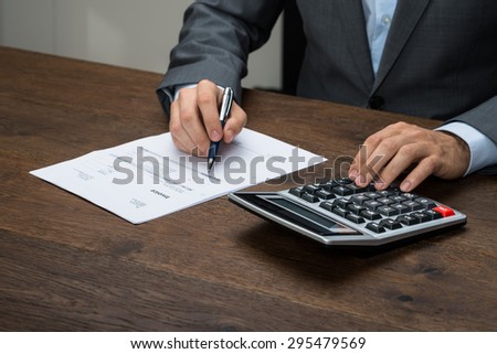 Close-up Of Businessman Calculating Invoice With Calculator At Desk