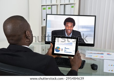 Businessman With Digital Tablet Video Chatting With Male Colleague On Computer In Office