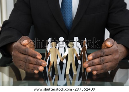 Close-up Of Businessman Hands Protecting Paper Cutout People At Desk