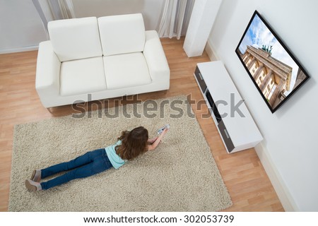 High Angle View Of Girl Lying On Carpet Watching Television At Home