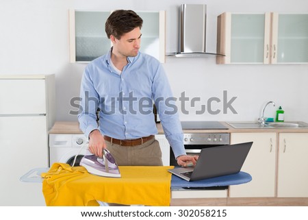 Young Man Typing On Laptop While Ironing Cloth In Kitchen