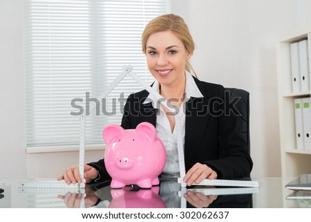 Happy Businesswoman Holding Ruler Folded In House Shape With Piggy Bank At Desk