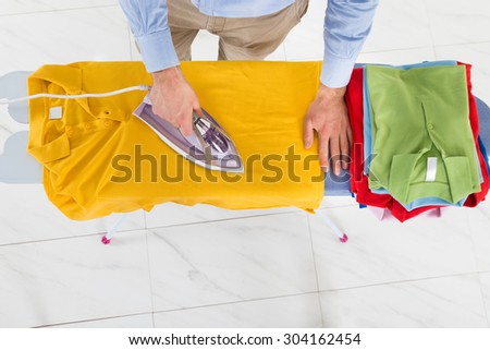 High Angle View Of Man Ironing Yellow T-shirt With Electric Iron On Ironing Board
