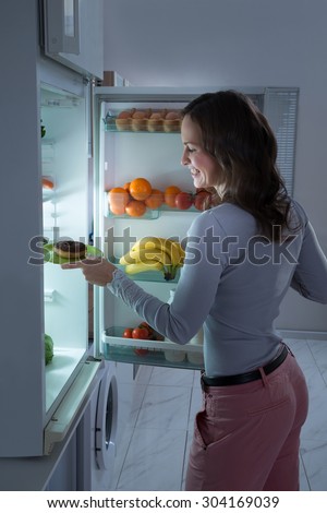 Happy Young Woman Taking Donut From Refrigerator At Night
