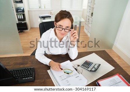 Young Businesswoman With Invoice And Magnifying Glass At Desk In Office