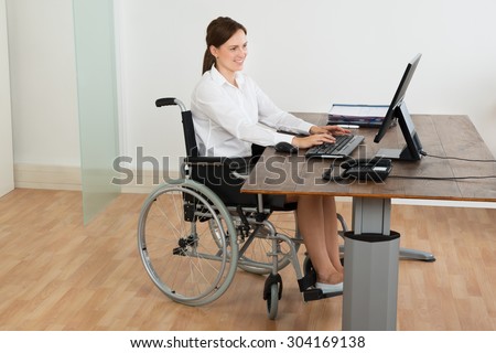 Happy Businesswoman Sitting On Wheelchair While Working On Computer At Desk