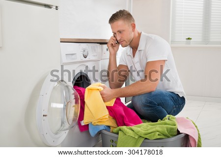 Young Man Talking On Mobile Phone While Putting Laundry In Washing Machine