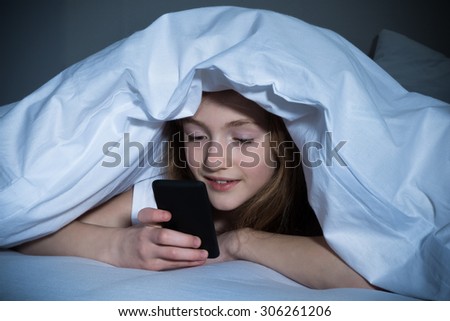Happy Girl Looking At Mobile Phone While Lying Under Blanket On The Bed