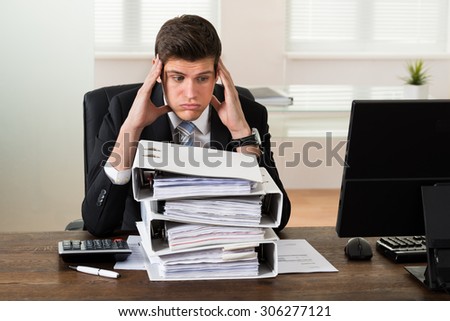 Stressed Young Businessman Looking At Stack Of Folders In Office