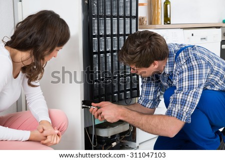 Young Repairman Repairing Refrigerator In Front Of Beautiful Woman