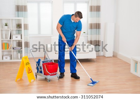 Happy Worker With Cleaning Equipments And Wet Floor Sign In House