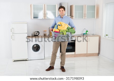 Young Handsome Man Carrying Basket With Heap Of Clothes In Kitchen