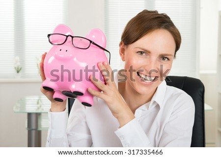 Portrait Of Happy Businesswoman Holding Piggybank With Eyeglasses In Office