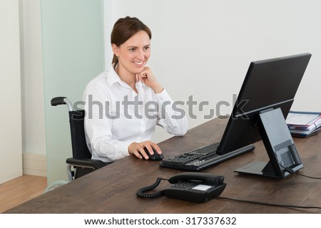 Happy Businesswoman Sitting On Wheelchair While Working On Computer At Desk