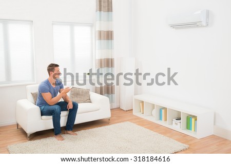 Young Man On Sofa Operating Air Conditioner With Remote Control