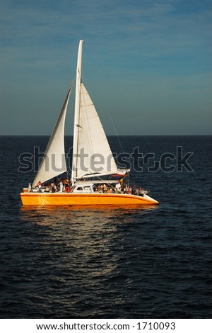 Catamaran with orange hull and white sail at sunset.  Deck is filled with people at a party.  There is a golden glow from sunset.  The hull and sails are reflected in the water.  There is a blue sky.