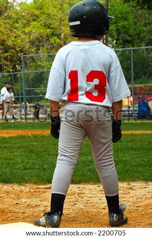 Young boy playing baseball.  He is standing on third  base.  He is wearing a helmut and his shirt says 13.  There is a player and coach visible on first base in the blurred background.